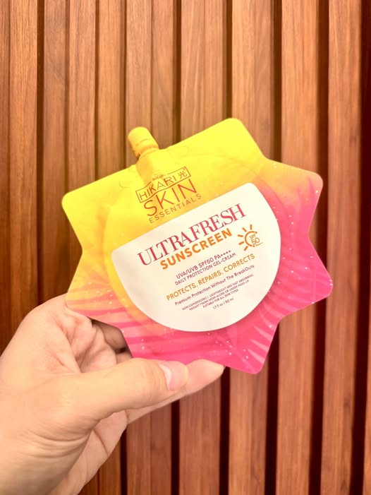 Hand holding a yellow and pink hikari skin ultrafresh sunscreen package with 'Hakuhodo Skin' branding against a wooden background.