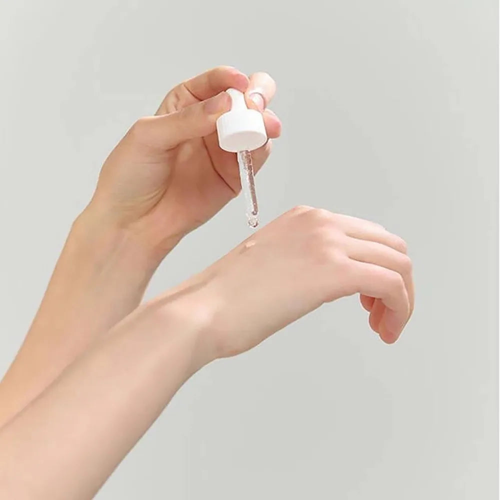 Hand holding a dropper with a clear liquid, applying it to another hand against a light gray background