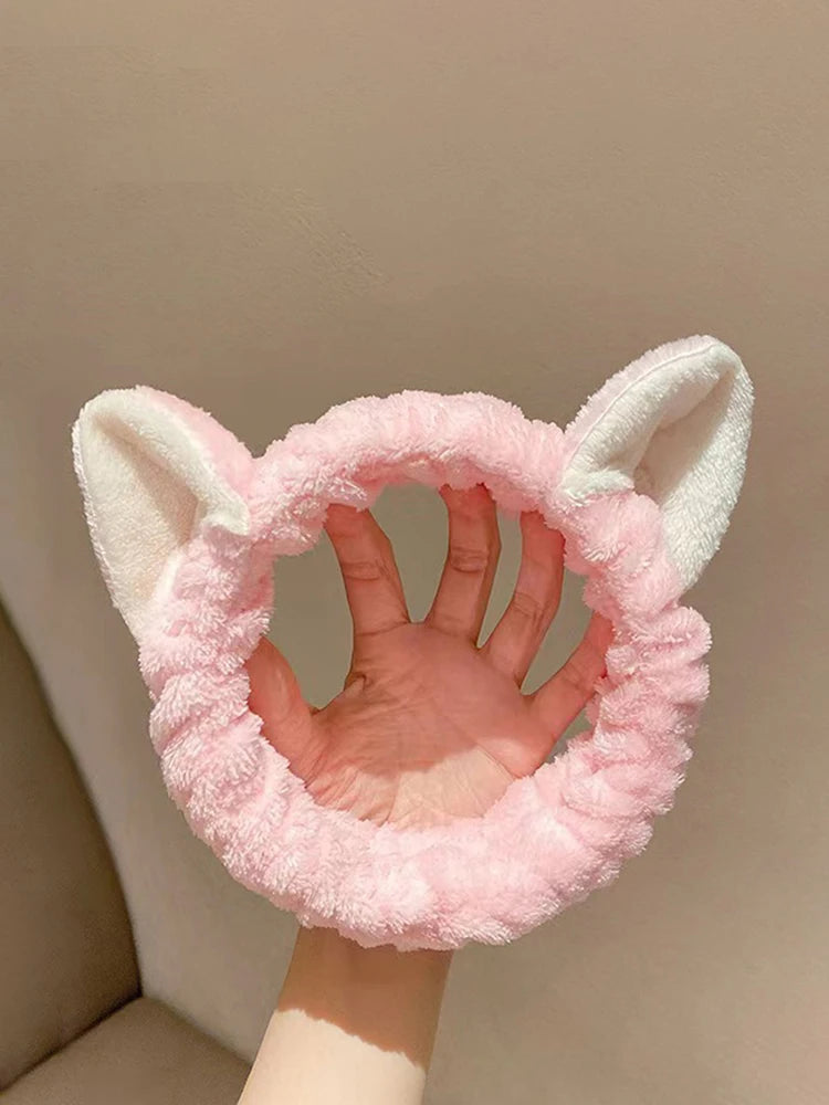 Pink fuzzy headband with cat ears held by a hand against a beige background